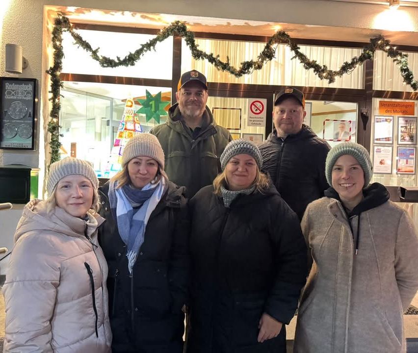 Eine Gruppe von sechs Erwachsenen, alle in Wintermänteln, lächeln und posieren für ein Foto vor einem Geschäft mit festlicher Dekoration.