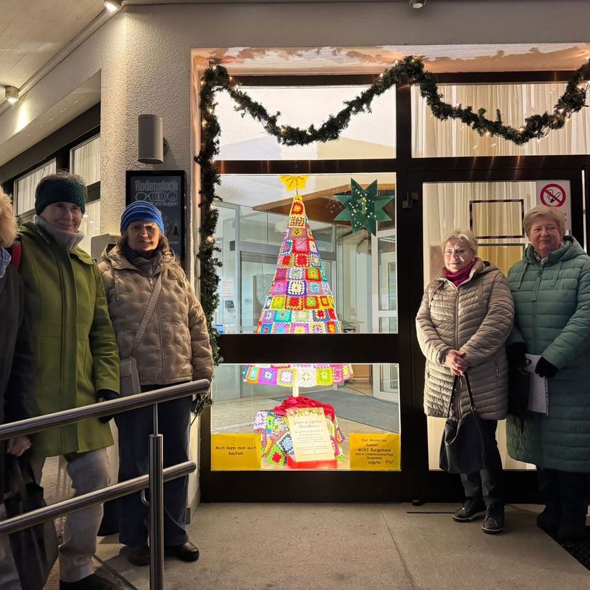 Fünf Frauen stehen vor einem Gebäude, lächeln und posieren für ein Foto. Der Eingang hat eine Weihnachtsbaumdekoration und ein Schild, das einen Verkauf anzeigt.