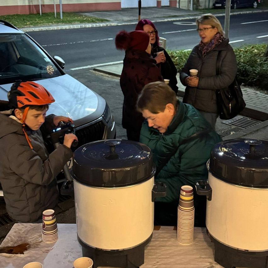 Eine Gruppe von Menschen versammelt sich um einen Tisch mit Kaffeekannen. Ein Junge im Helm hält eine Kamera. Ein Mann lächelt, und andere stehen in der Nähe mit Tassen. Ein Auto ist am Straßenrand geparkt.