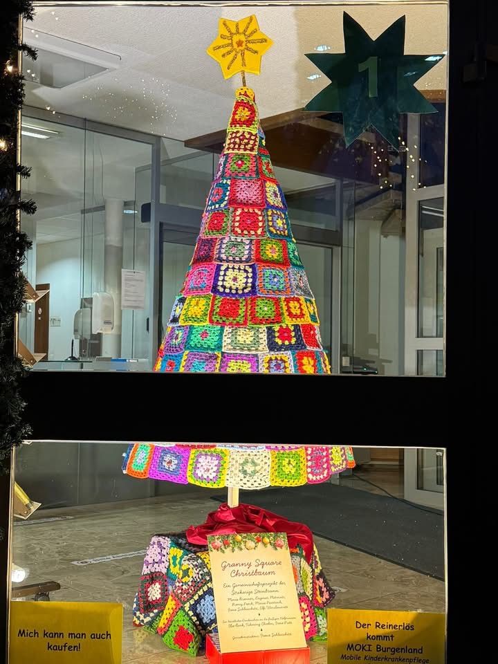 Ein farbenfroher gehäkelter Weihnachtsbaum steht in einer Schaufensterscheibe mit Reflexionen eines Innenbüroraums und eines großen Schildes. Der Baum hat einen Stern obenauf und steht auf einem dekorativ gestalteten Sockel.