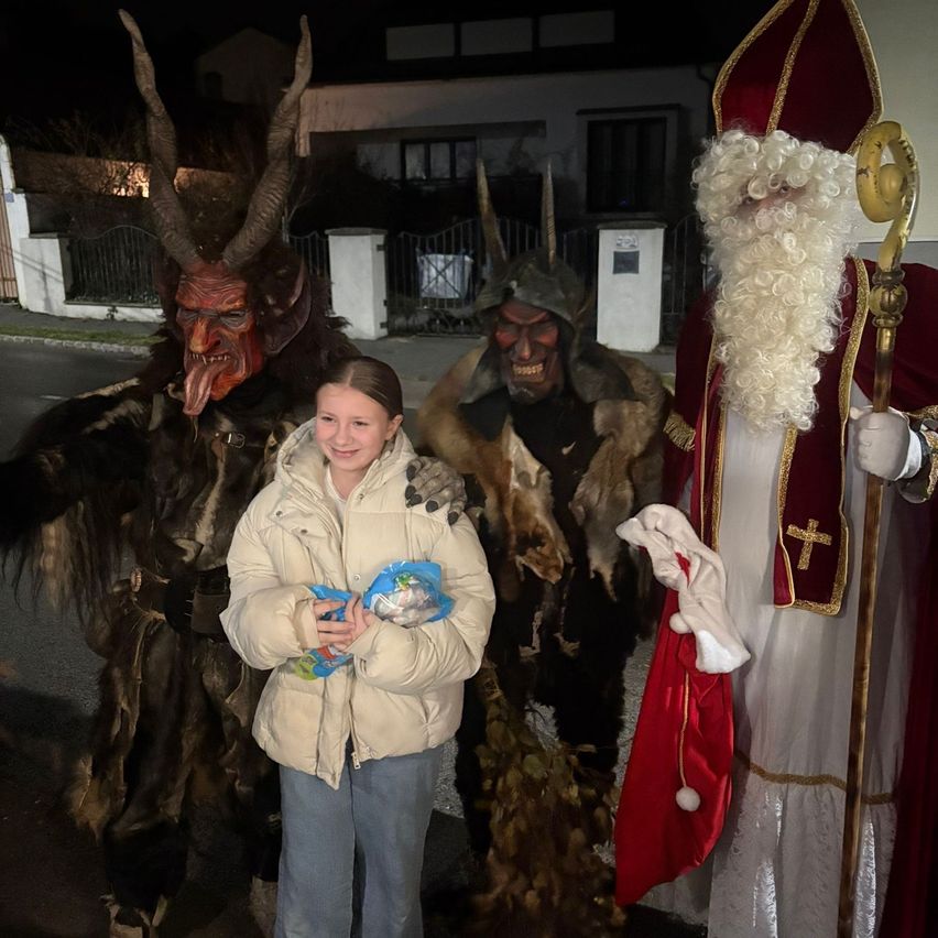 Ein junges Mädchen posiert mit einer Person in Teufelskostüm und einer anderen in Santa Claus Kostüm, beide in Fell gekleidet.