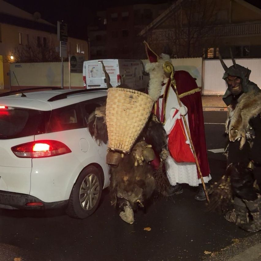 Zwei Menschen in Kostümen stehen auf einer Straße mit einem weißen Auto, einer trägt einen Korb und der andere einen Stab. Dahinter ist ein Lieferwagen geparkt.