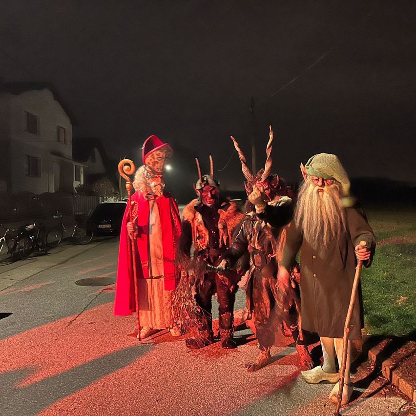 Fünf Personen in aufwendigen Kostümen, möglicherweise für eine Halloween-Veranstaltung, stehen bei Nacht auf der Straße. Die Kostüme umfassen einen roten Umhang, eine gehörnte Maske und einen langen Stab. Im Hintergrund ist ein Haus mit mehreren Fenstern zu sehen.