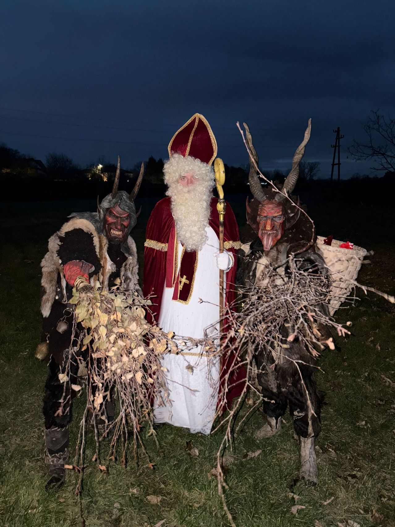 Ein Mann in der Kleidung des Heiligen Nikolaus steht zwischen zwei Männern in Teufelskostümen, alle halten Stöcke. Die Szene spielt bei Nacht mit einem dunklen Himmel.