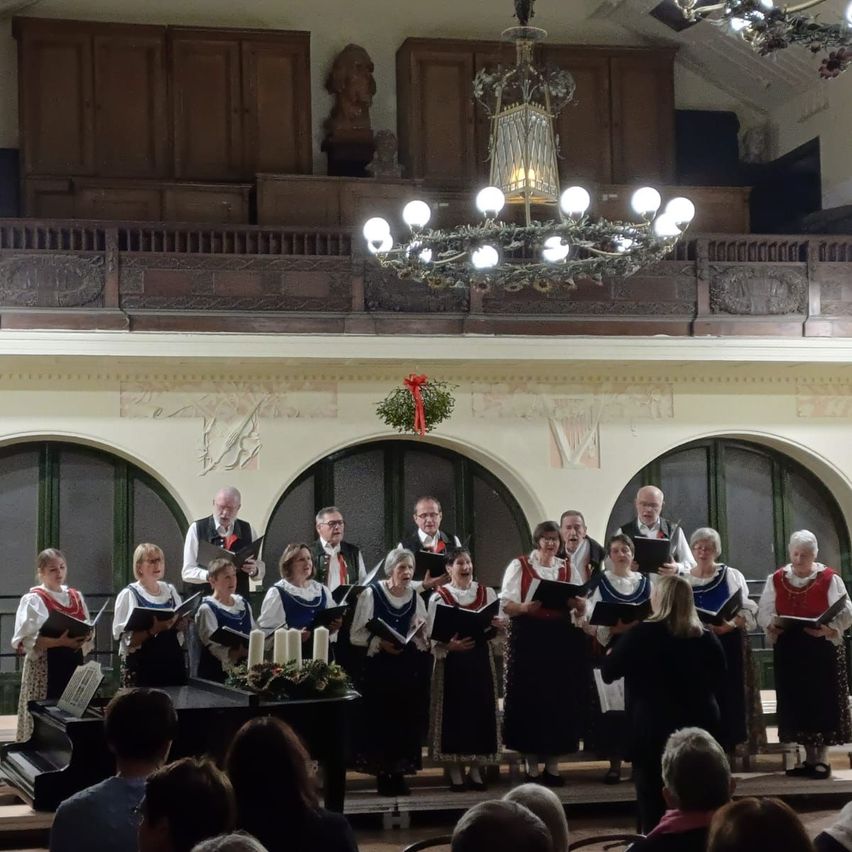 Ein Chor in traditioneller Kleidung steht auf der Bühne, hält Notenblätter in der Hand, mit einem Pianisten und einer Laterne darüber und einer Weihnachtsdekoration. Ein Publikum sieht von vorne zu.