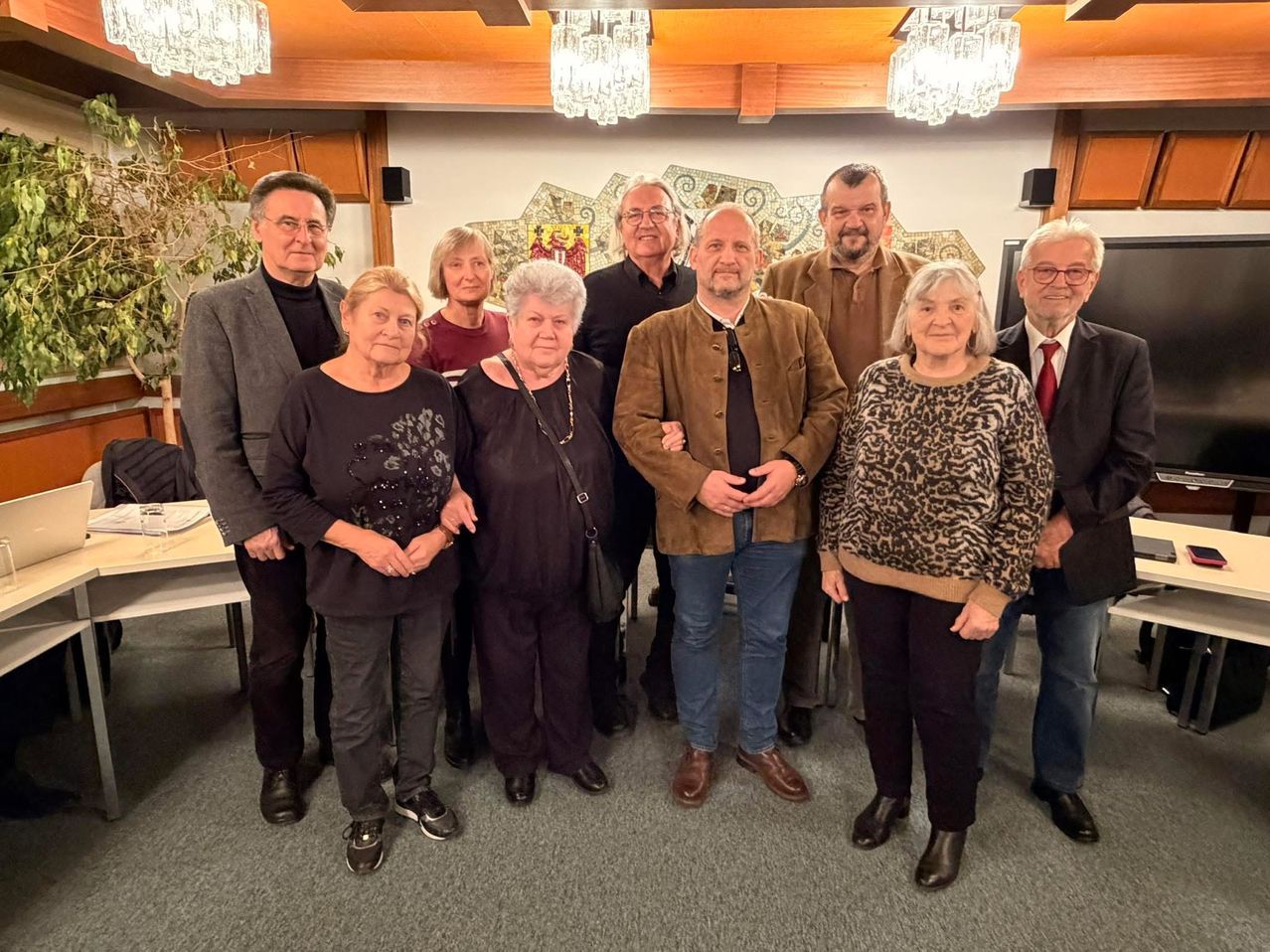 Eine Gruppe von Menschen steht in einem Raum und posiert möglicherweise für ein Foto. Sie sind lässig gekleidet. Hinter ihnen befindet sich eine Wand mit einem dekorativen Fliesenmuster. Auf der linken Seite steht ein Tisch mit einer Pflanze, und Kronleuchter hängen von der Decke.