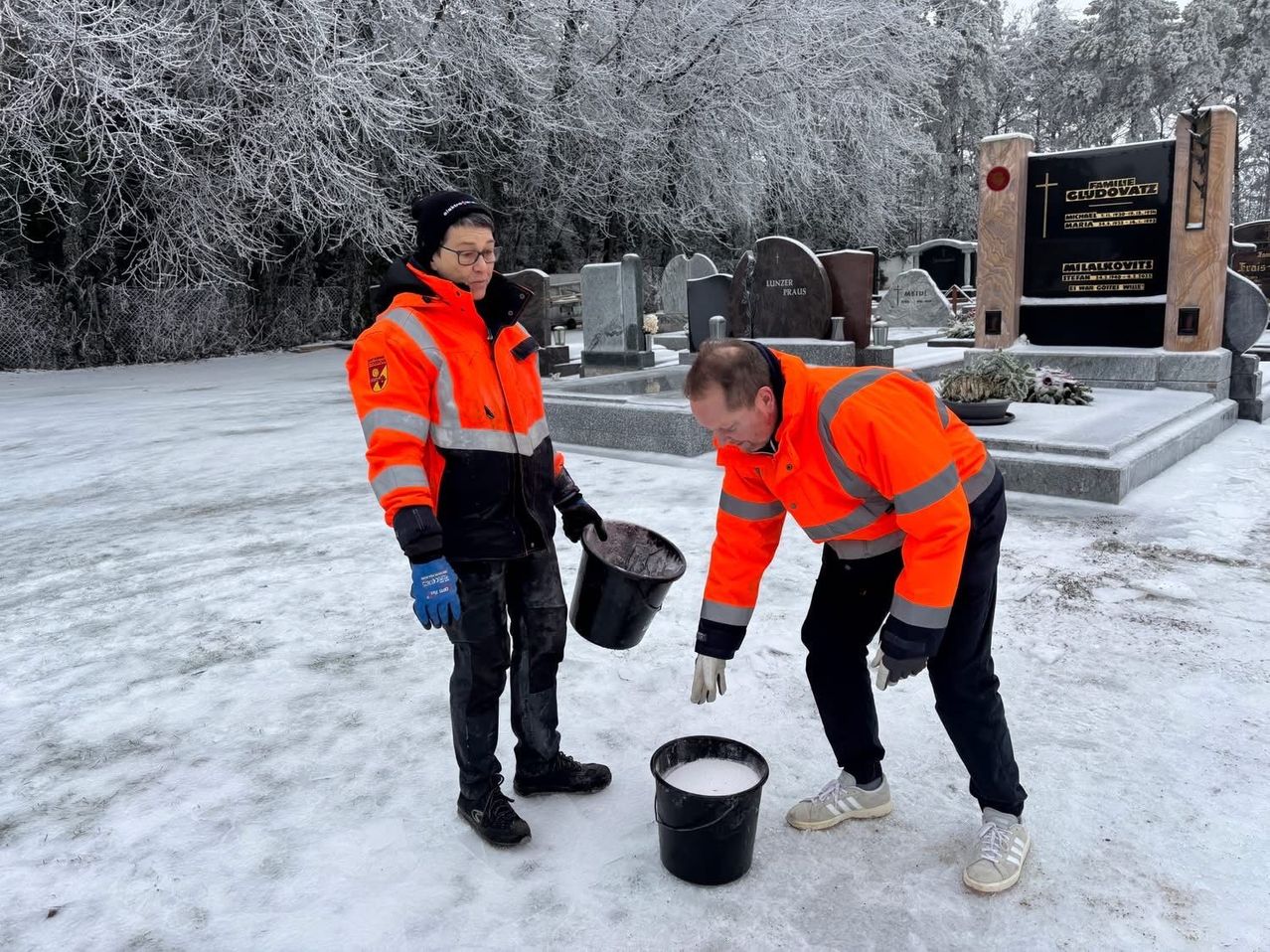 Zwei Arbeiter in orangefarbenen Jacken und Handschuhen gießen einen Eimer Flüssigkeit in einen schneebedeckten Friedhof.