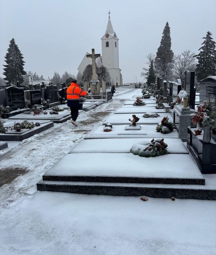 Eine Person in einer orangefarbenen Warnweste geht in einem verschneiten Friedhof und reinigt Grabstätten. Ein Kirchengebäude mit einem Turm ist im Hintergrund zu sehen.