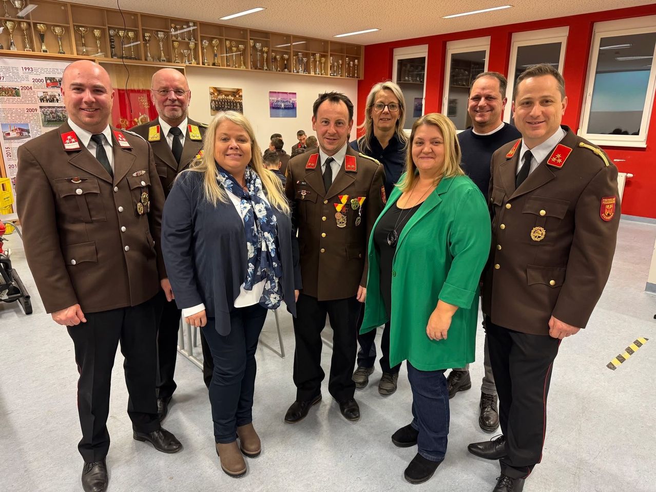 Eine Gruppe von Erwachsenen, darunter Männer und Frauen in Militäruniformen und andere in Freizeitkleidung, stehen in einem Raum mit roten Wänden und Trophäenregalen.