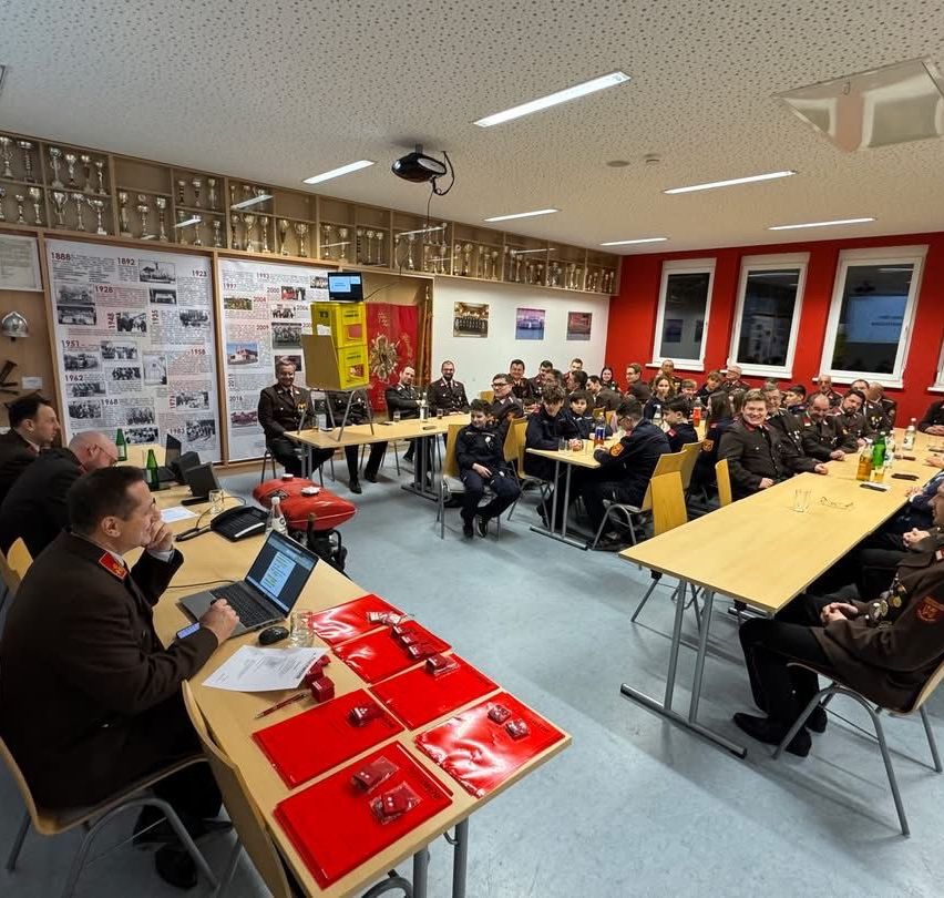 Ein Besprechungsraum mit roten Wänden und mehreren Tischen, wo Personen in Uniformen versammelt sind. Ein Mann vorne sitzt, während andere stehen und sitzen. Laptops, Flaschen und rote Schachteln sind auf den Tischen.
