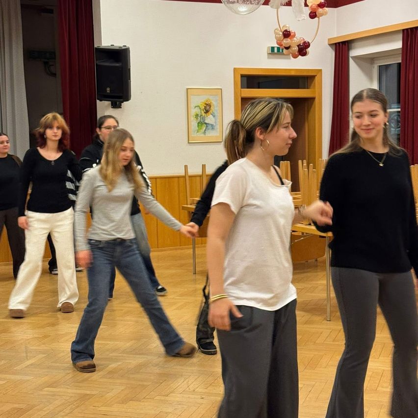 Eine Gruppe von Frauen tanzt in einem Raum. Einige tragen Turnschuhe, andere Stiefel. Sie sind alle lässig gekleidet. Hinter ihnen ist ein Lautsprecher an der Wand montiert.