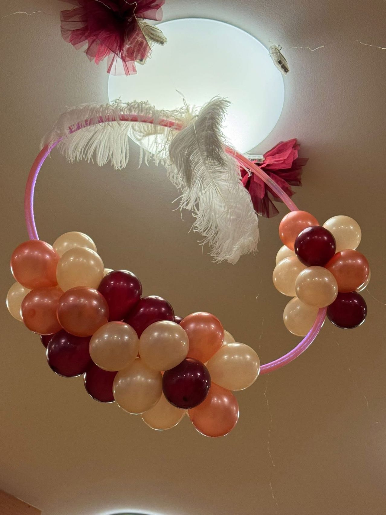 Ein rosafarbener Ballonbogen-Dekor mit roten und pfirsichfarbenen Ballons, einer weißen Feder und einem roten Band. Der Bogen ist an einer weißen Leuchte an der Decke befestigt.