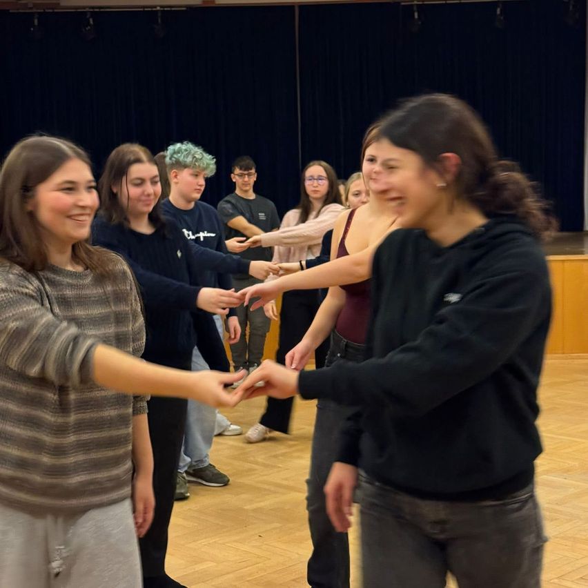 Eine Gruppe junger Menschen tanzt zusammen auf einem Holzboden. Einige lächeln und halten sich an den Händen, während andere zusehen.