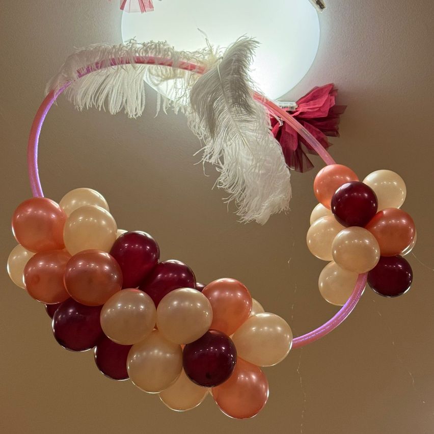 Ein rosafarbener Ballonbogen mit weißen Federn und roten Bändern hängt von der Decke. Der Bogen ist mit einer Mischung aus roten, pfirsichfarbenen und weißen Ballons gefüllt.
