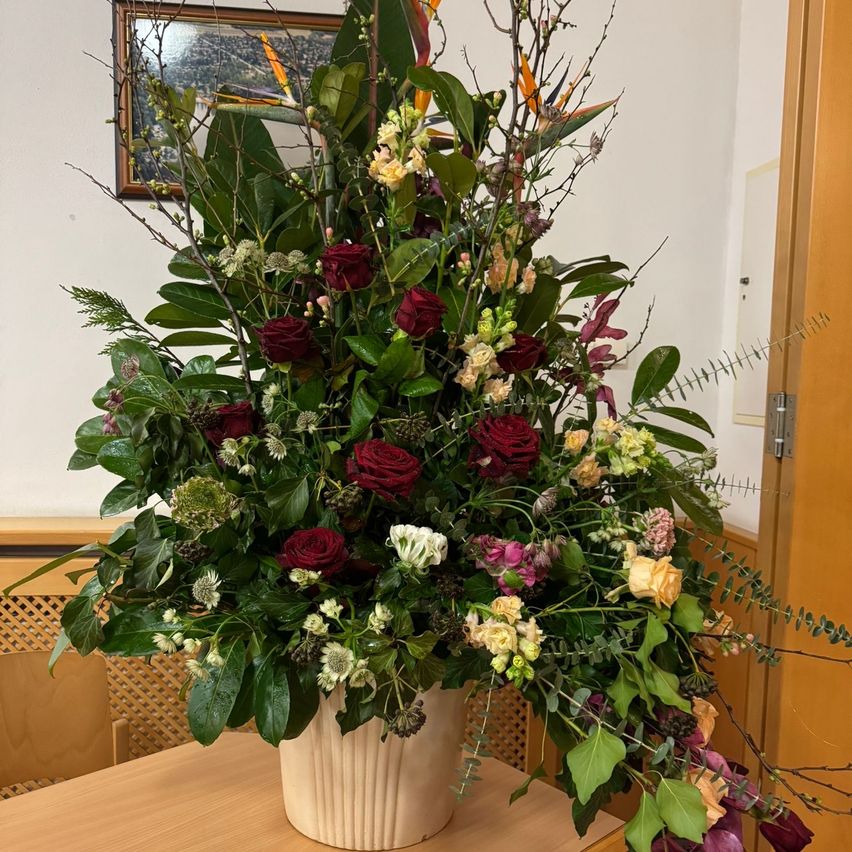 Eine große Blumenarrangement mit einer Mischung aus roten, weißen und gelben Blumen in einem hohen weißen Vasen, auf einem holznen Tisch in einem Raum mit einer weißen Wand und einem gerahmten Bild.
