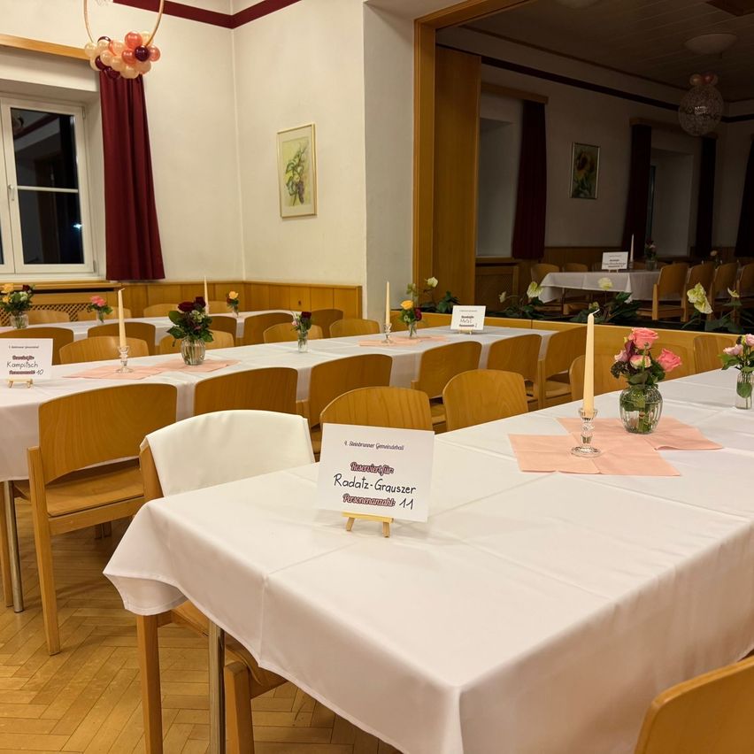 Ein Esszimmer, das für einen besonderen Anlass vorbereitet wurde, mit langen Tischen, die mit weißen Tischdecken bedeckt sind. Jeder Tisch hat ein Schild mit den Namen der Gäste. Die Stühle sind ordentlich angeordnet, und Blumenarrangements sind auf den Tischen platziert. Der Raum ist gut beleuchtet mit natürlichem Licht aus den Fenstern.