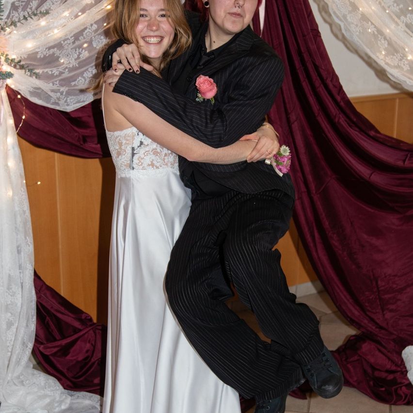 Ein Paar, die Frau in einem weißen Kleid und der Mann im schwarzen Anzug, teilen einen freudigen Moment mit einem Blumenstrauß.