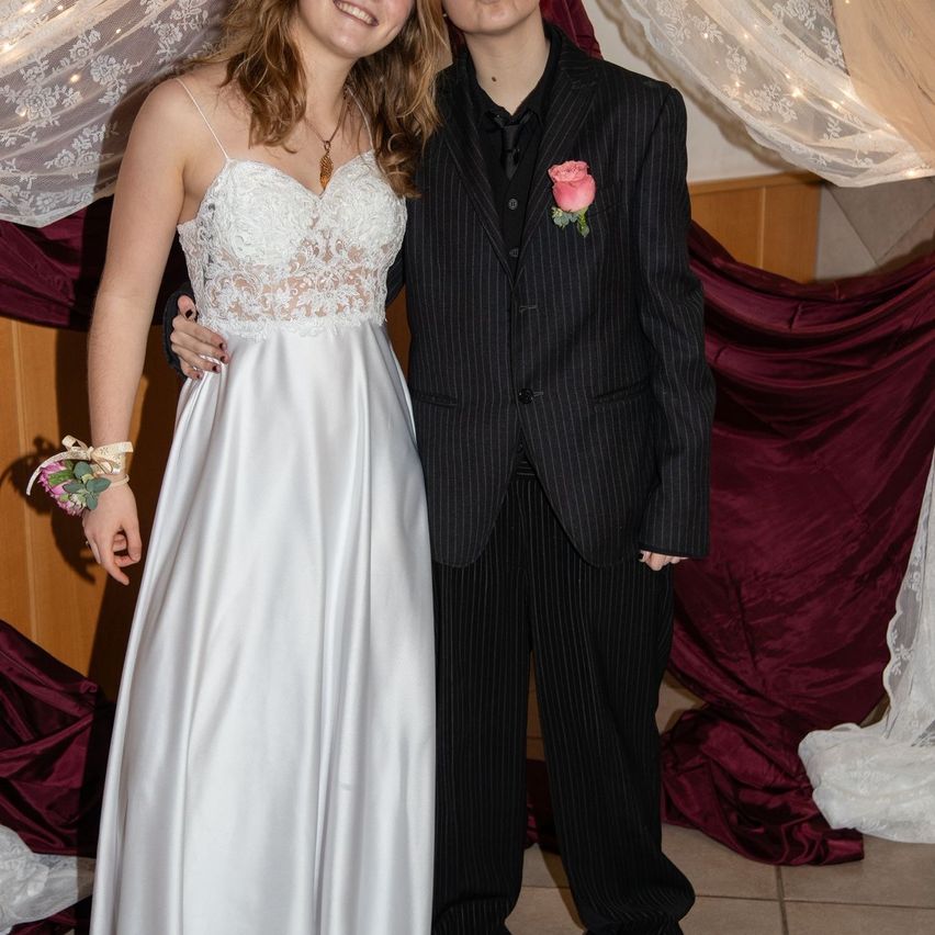 Ein junges Paar steht zusammen, die Frau in einem weißen Kleid und der Mann in einem schwarzen Anzug mit einer rosa Rose im Revers. Sie posieren für ein Foto vor einem Hintergrund aus roten und weißen Vorhängen.