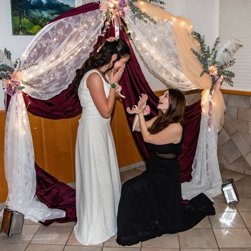 Zwei Frauen, eine in einem weißen Kleid und die andere in einem schwarzen Kleid, knien unter einem geschmückten Bogen. Die Frau im weißen Kleid hält eine Blume, und die Frau im schwarzen Kleid lächelt sie an. Ein Gemälde hängt an der Wand hinter ihnen.