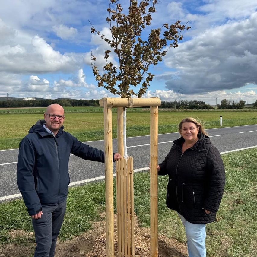 Zwei Personen stehen neben einem jungen Baum in einem Feld und lächeln. Eine Straße verläuft hinter ihnen, mit Wolken am Himmel.
