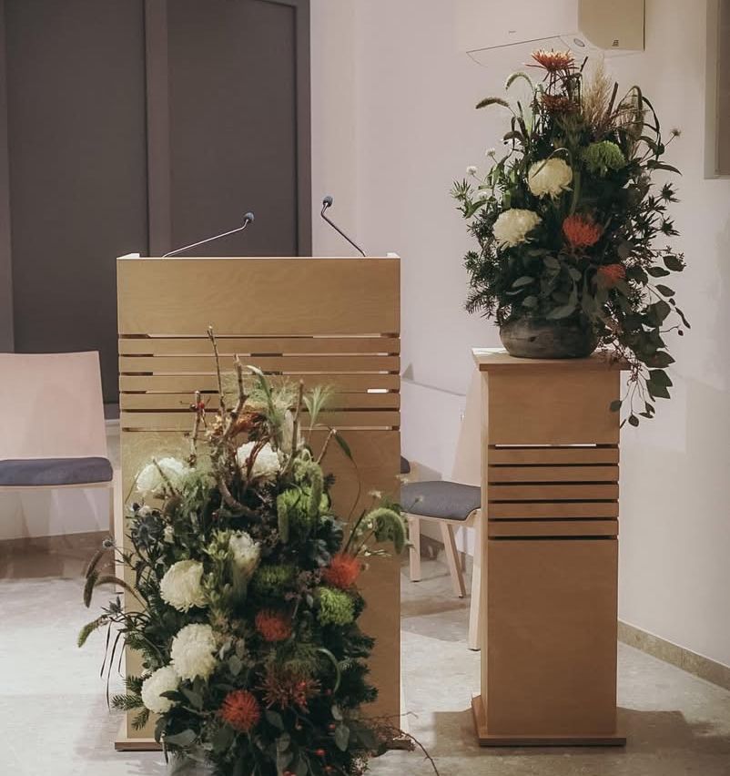 A wooden podium with a microphone is placed next to a wooden stand with a flower arrangement. The room has a white wall and gray floor.