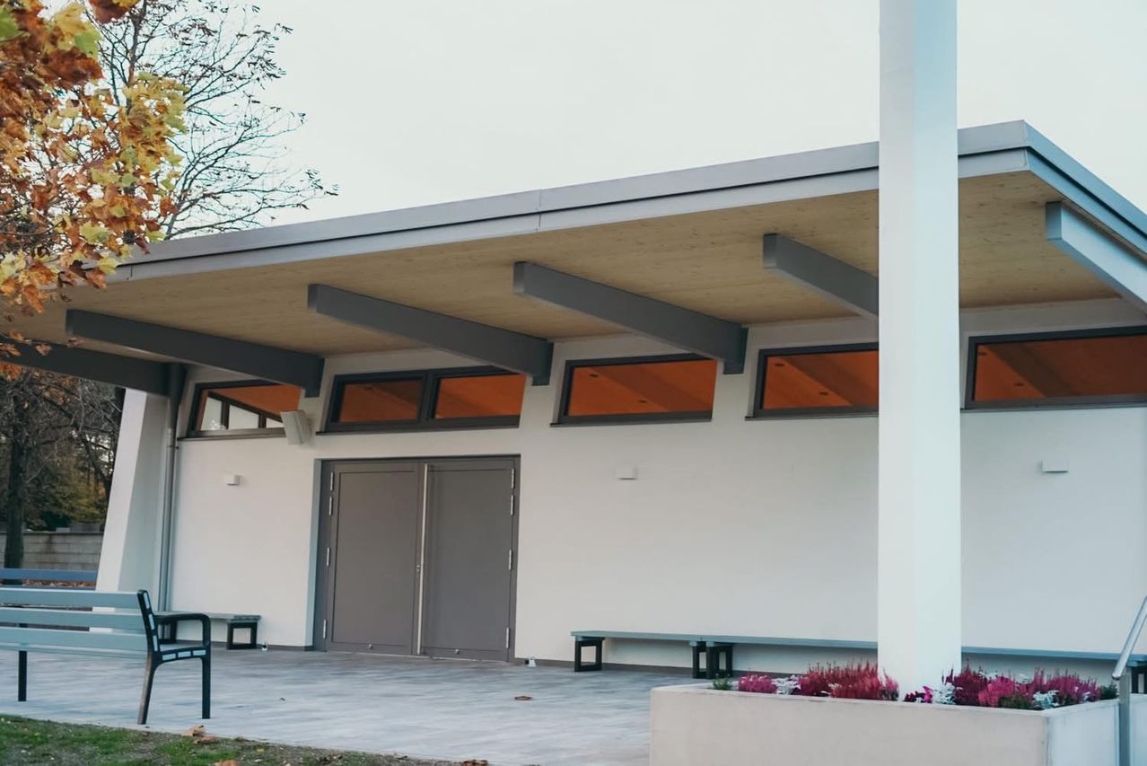 A modern building exterior with a double gray door, gray beams, and orange windows. There are two benches on either side of the entrance.