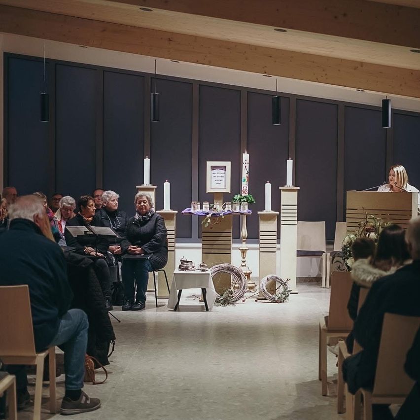 A woman speaks at a podium in a chapel. People sit in chairs around a table with lit candles. Wooden beams and blue drapes adorn the walls.