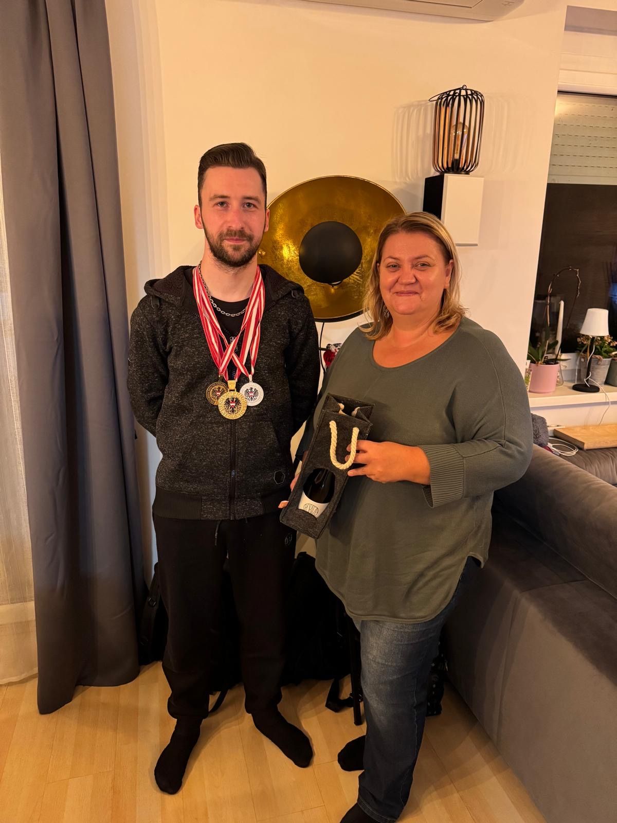 A man with medals and a woman with a gift bag stand in a room with a couch, lamp, and curtain.