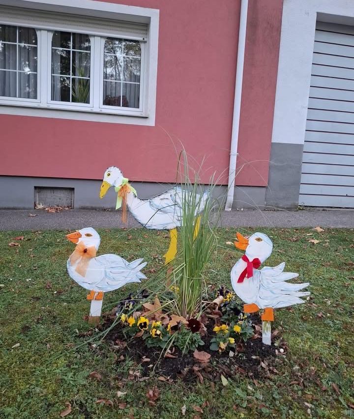 Three decorative ducks stand in a garden with yellow and red ribbons. One is a mother duck, and the others are her ducklings.