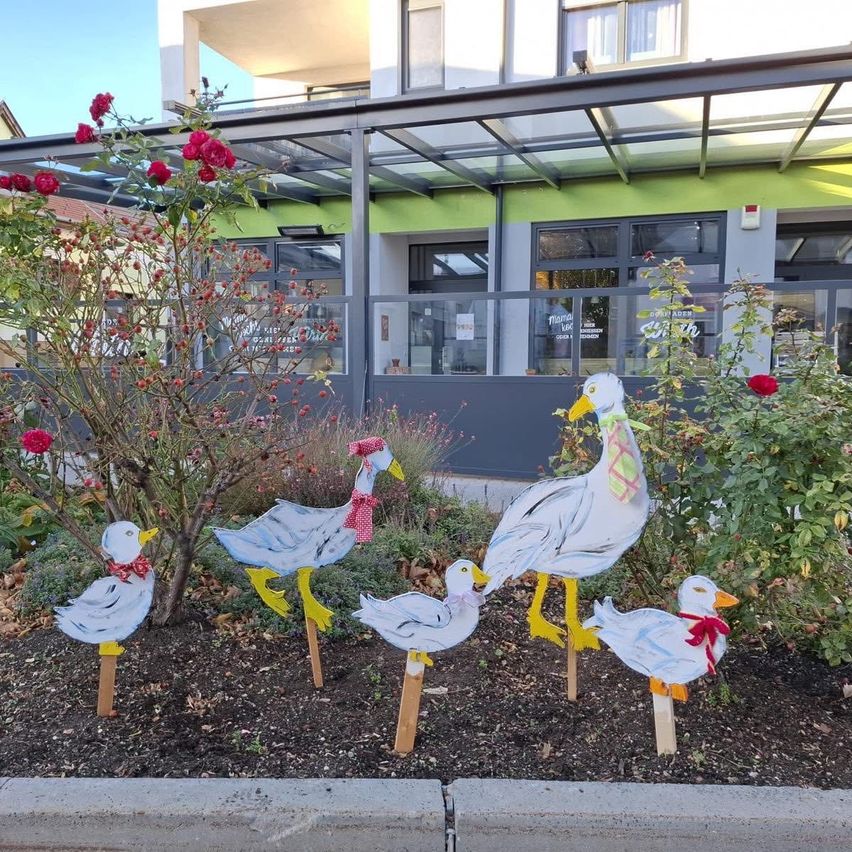 A row of painted duck sculptures with red bows stands in a garden with red flowers and plants in front of a building.