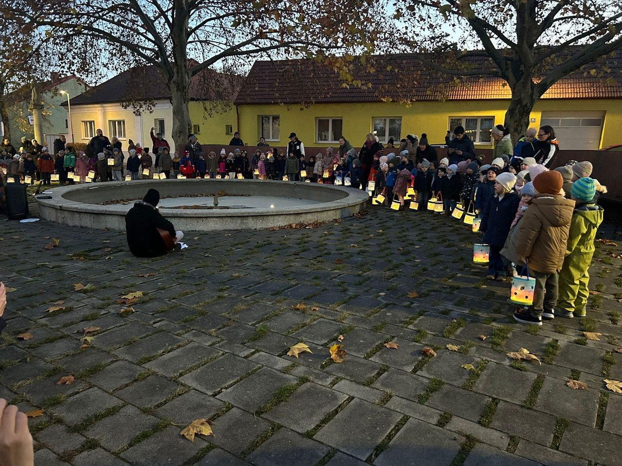 Many children holding glowing lanterns form a circle around a fountain, a person sits in the center. The scene is set in a courtyard with yellow buildings and trees.