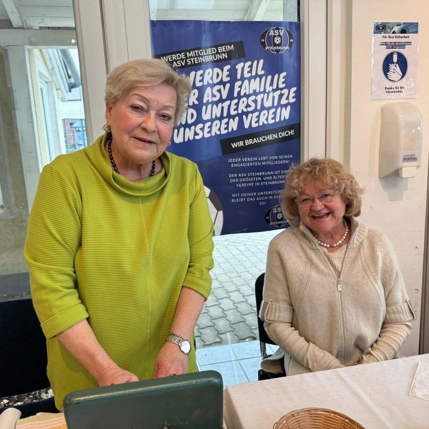 Zwei Frauen, die lächelnd vor einem Banner posieren. Eine Frau in Grün, die andere in Beige. Ein Seifenspender und ein Schild befinden sich an der Wand hinter ihnen.