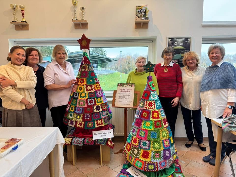 Eine Gruppe von Frauen steht um zwei gehäkelte Weihnachtsbäume in einem Raum. Ein Tisch und ein weißes Tuch sind im Vordergrund.