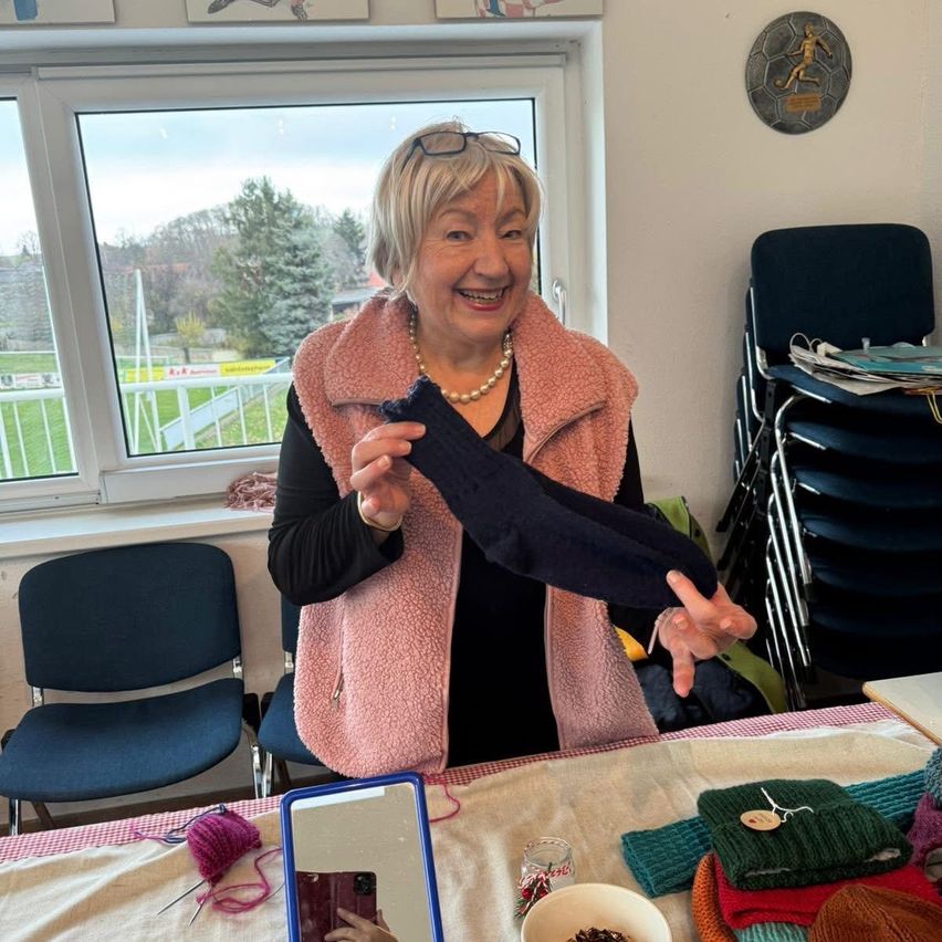Eine lächelnde Frau in einem rosa Weste und schwarzem Oberteil hält einen blauen gestrickten Gegenstand, sitzt an einem Tisch mit Strickzubehör. Hinter ihr zeigt ein Fenster Bäume und ein grasiges Feld.