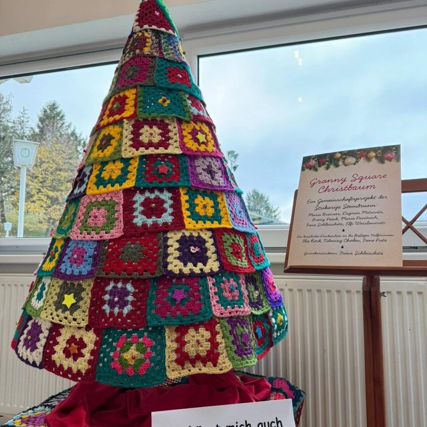 Ein gehäkelter Weihnachtsbaum aus bunten Quadraten steht vor einem Fenster mit Blick auf Bäume. Ein Schild mit der Aufschrift 'Granny Square Christmas Tree' und einer Beschreibung. Ein weißes Schild mit schwarzem Text liegt auf einem roten Tuch unter dem Baum.