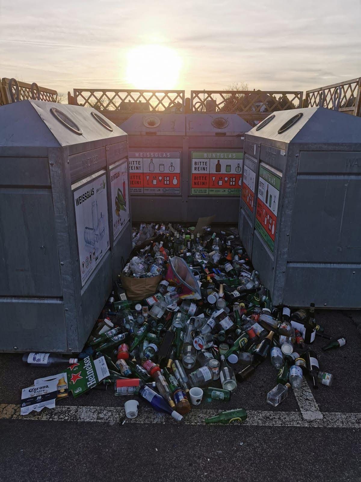Ein Foto von einem Haufen leerer Flaschen und Dosen vor Recyclingbehältern. Die Flaschen und Dosen liegen verstreut auf dem Boden.