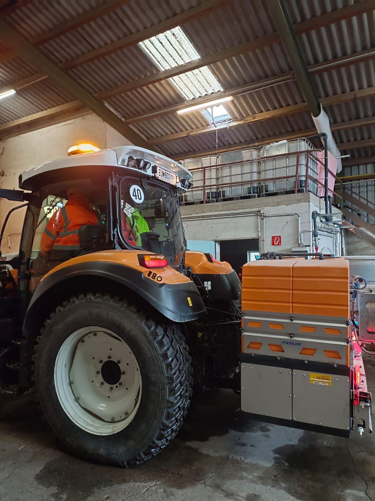 Ein orangefarbener Traktor mit der Nummer 40 steht in einem großen Gebäude. Der Traktor hat einen großen Anbau am Heck, möglicherweise zum Transport von Ausrüstung. Das Innere des Gebäudes besteht aus Metallstrukturen und großen Tanks.