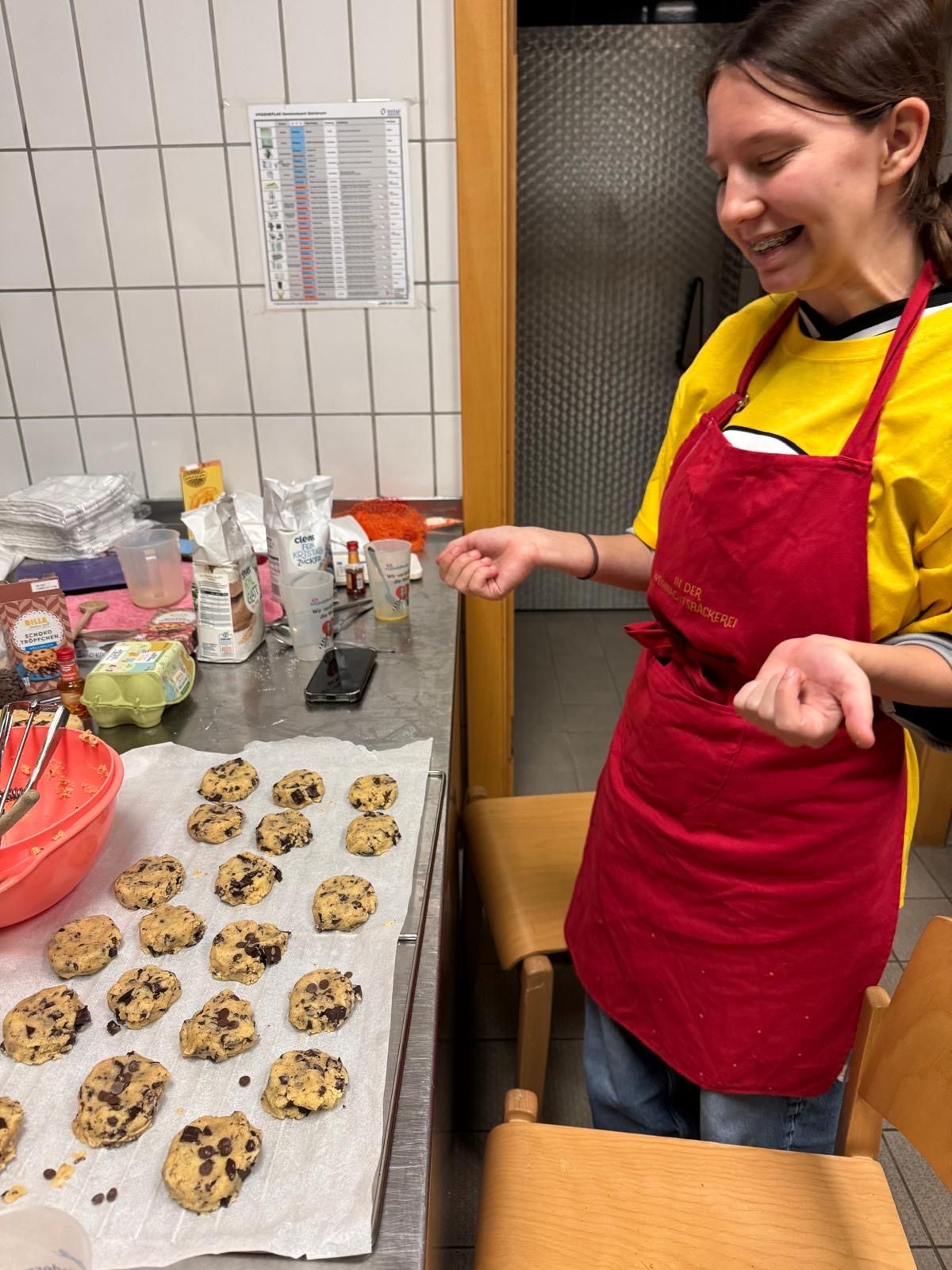 Eine Frau in einem roten Schürzen steht in einer Küche, lächelt und zeigt auf Kekse auf einem Backblech. Der Tisch hat eine Schüssel, Messbecher und verschiedene Zutaten.