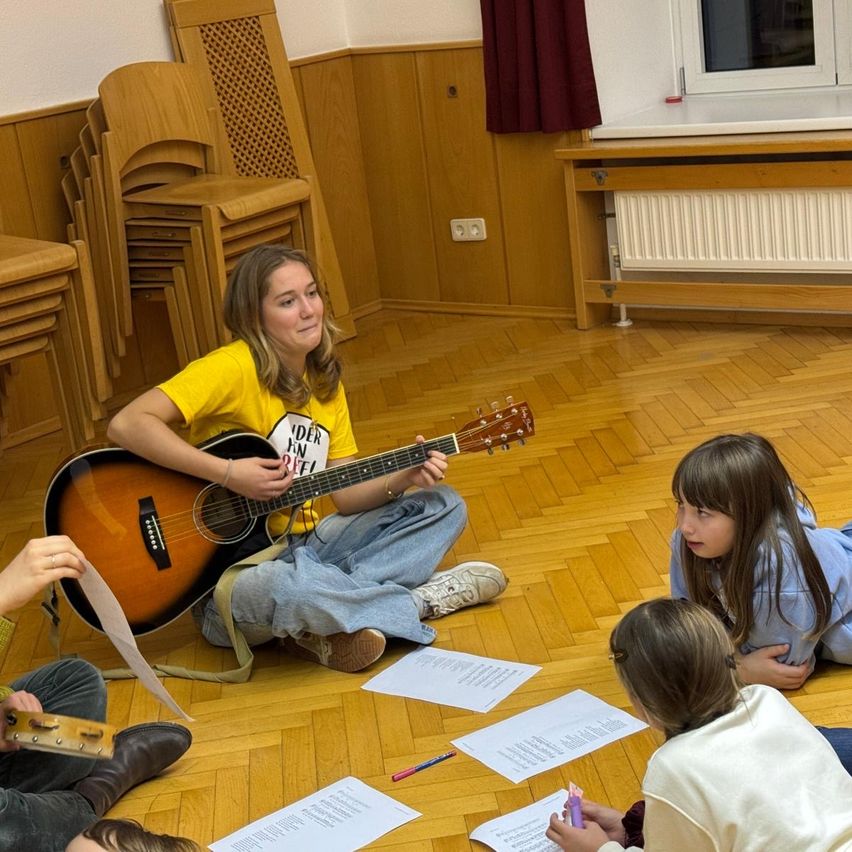 Eine Frau sitzt auf dem Boden und spielt Gitarre, während drei Kinder um sie herumsitzen und sie anschauen. Papiere sind auf dem Boden verstreut. Hinter ihr ist ein Heizkörper an der Wand montiert.