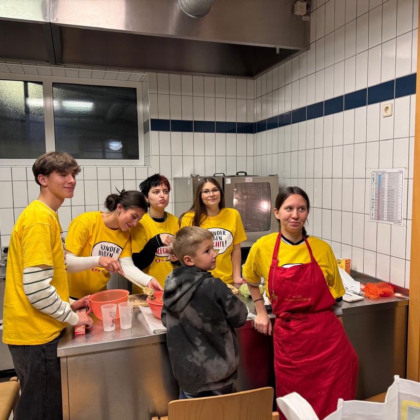 Eine Gruppe von Menschen in gelben T-Shirts bereitet Essen in einer Küche zu. Eine Frau in einer roten Schürze steht vor einem Tresen mit einem jungen Jungen davor. Es gibt Schüsseln, Becher und einen Beutel auf dem Tresen. Dahinter befindet sich ein Fenster und eine Wand mit Fliesen.