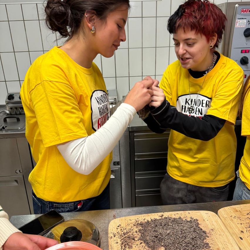 Zwei Frauen in gelben T-Shirts stehen in einer Küche, eine hält die Hand der anderen und beide lächeln. Sie stehen vor einem Tisch mit Schneidebrettern, und ein Handy und ein Topfdeckel sind in der Nähe.