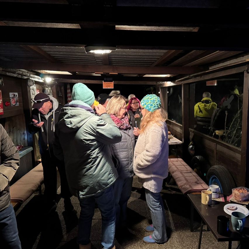 Eine Gruppe von Menschen steht in einem schwach beleuchteten Raum. Sie tragen Winterkleidung und einige tragen Mützen. Ein Tisch mit Tassen und Tellern ist in der Nähe.