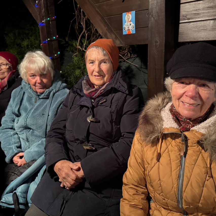 Drei Frauen in Wintermänteln sitzen nebeneinander in einem hölzernen Pavillon mit Weihnachtslichtern.