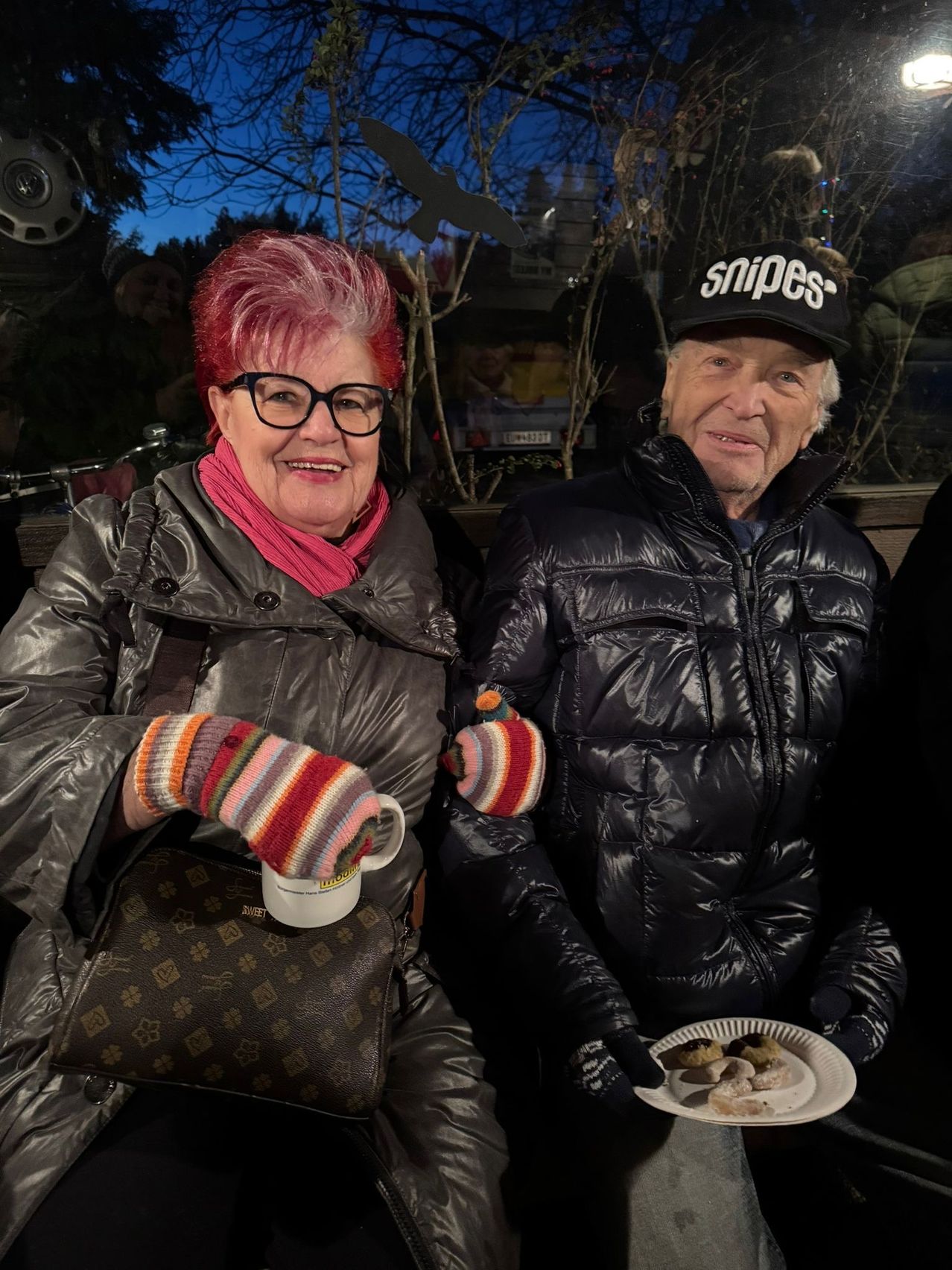 Ein älteres Paar sitzt zusammen und lächelt für die Kamera. Die Frau hat rote Haare und trägt eine Brille und einen Schal. Der Mann trägt einen schwarzen Hut und Handschuhe und hält einen Teller mit Essen. Sie sind für kaltes Wetter gekleidet.