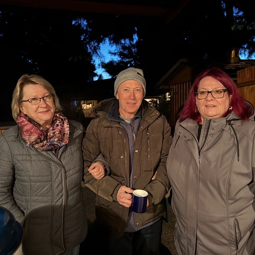 Drei Personen stehen eng beieinander und lächeln in die Kamera. Der Mann in der Mitte hält eine blaue Tasse. Sie sind in Winterkleidung gekleidet. Dahinter befinden sich Bäume und ein Holzzaun.