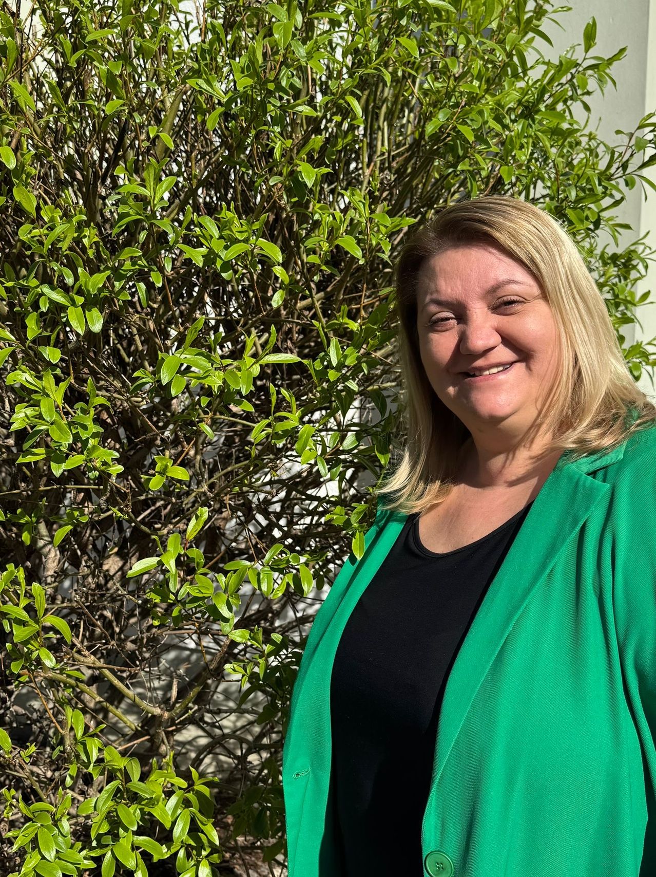 Eine Frau mit blondem Haar lächelt und steht vor einem Busch mit grünen Blättern. Sie trägt eine grüne Jacke und ein schwarzes Oberteil. Der Hintergrund ist unscharf.