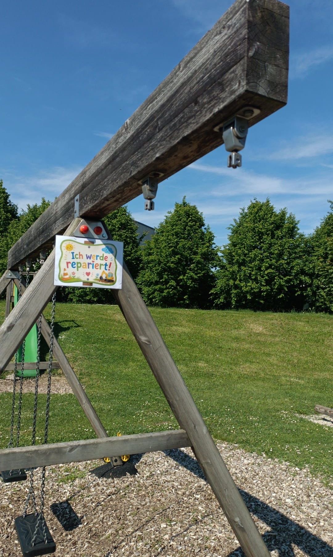 A wooden swing set with a sign saying 'I would repair!' hangs over a grassy area with trees.