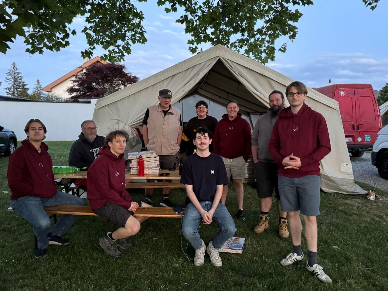 A group of people in maroon and black hoodies and t-shirts stand and sit in front of a tent with a table and books.
