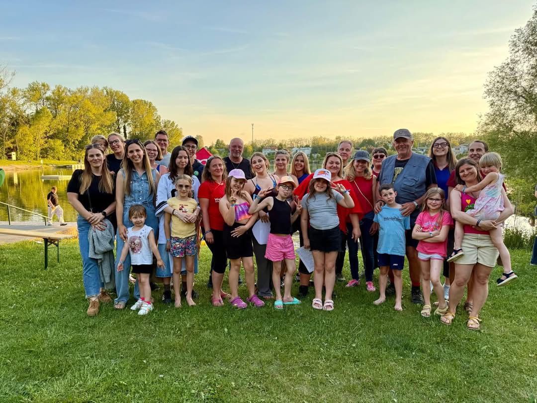 A large group of people, including children, pose for a photo in a grassy area during the day. Trees and a clear sky are in the background.