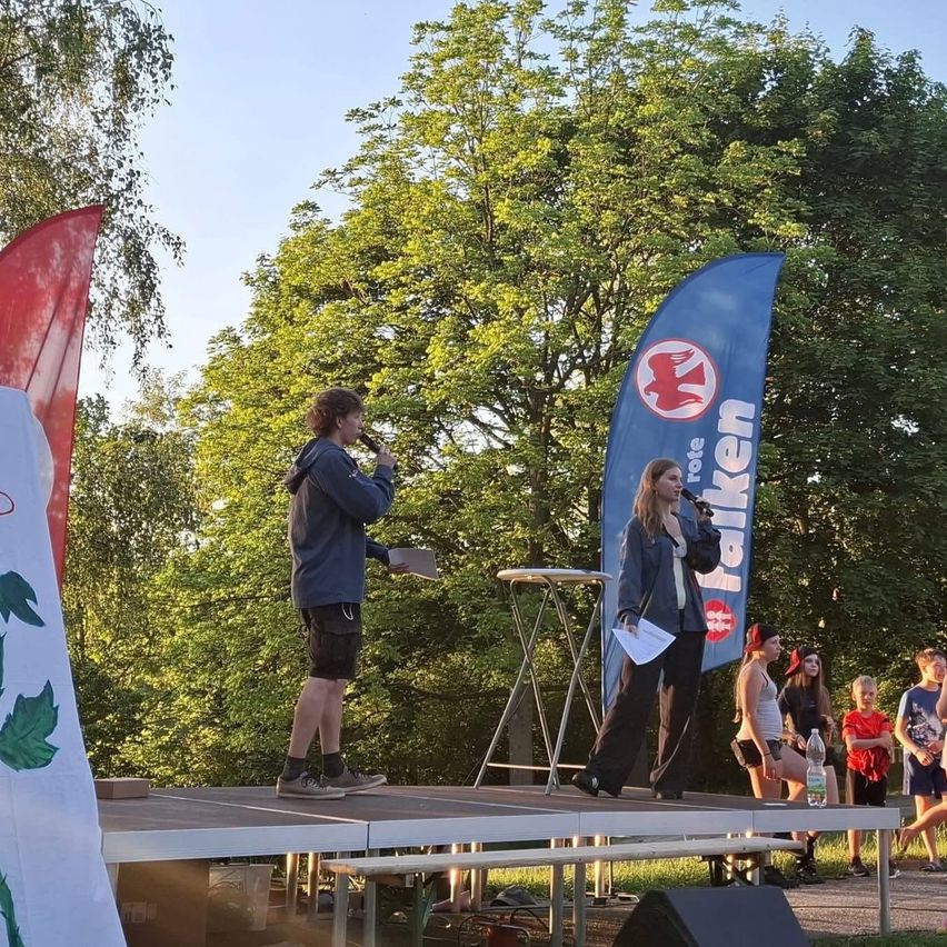 Two individuals stand on stage with microphones, one holding papers. A blue banner and a red flag are behind them. Several people sit and stand behind the stage. Trees are in the background.